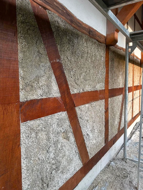 Photographie d'un artisan en train de poser de l'isolation en chanvre ou paille sur la paroi intérieure d'un bâtiment ancien, concentré sur son travail, portant des vêtements de travail appropriés et des équipements de sécurité. L'ambiance doit montrer la rigueur technique et le savoir-faire artisanal, avec les matériaux biosourcés visibles et identifiables.