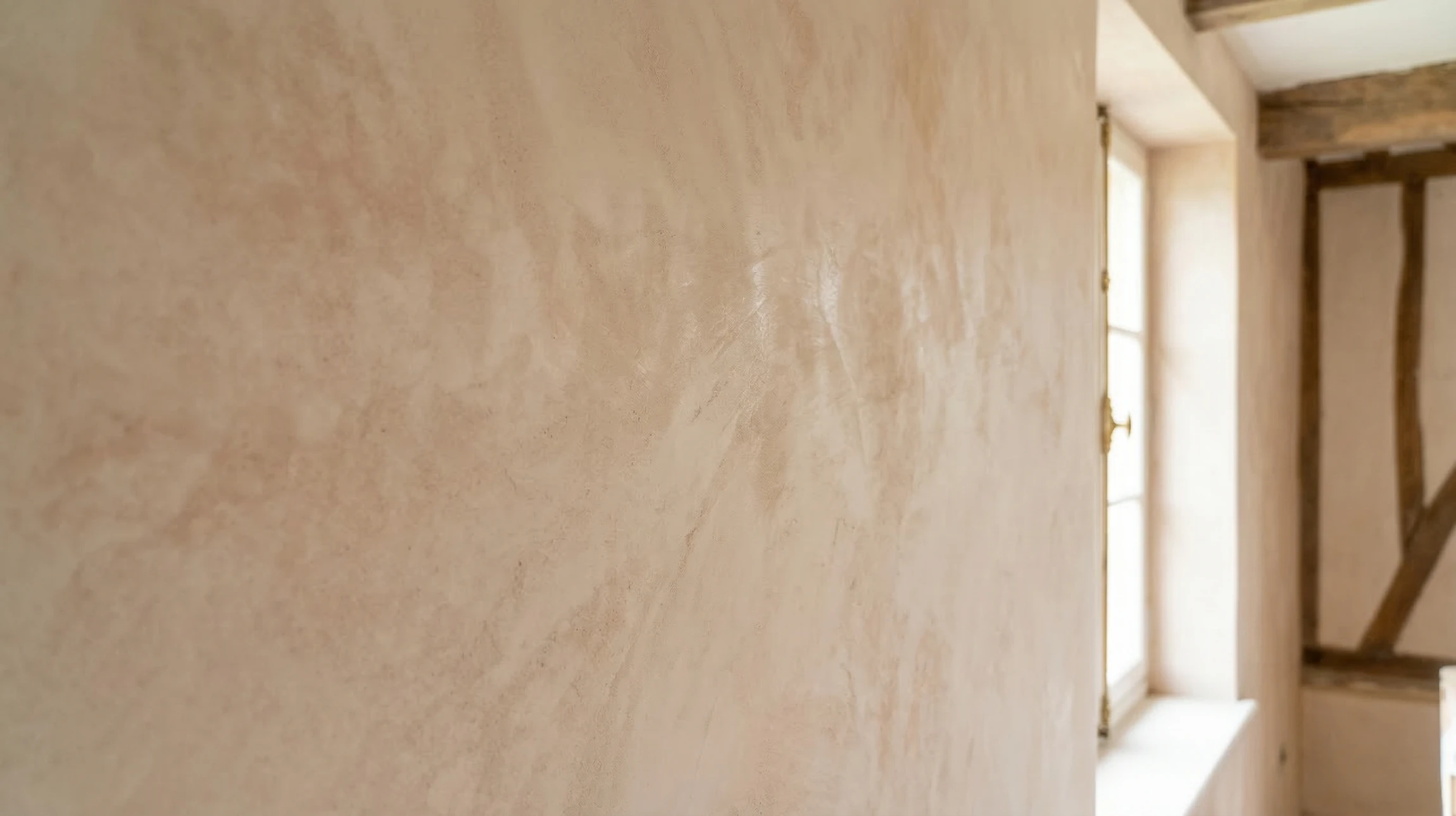 Gros plan détaillé sur un mur ancien en cours de rénovation avec application d'enduit naturel à base de chaux et terre. La surface murale montre partiellement l'enduit blanc/beige clair fraîchement appliqué au spatule en cours de lissage, avec des traces de travail à la main visibles. Le reste du mur présente l'état ancien rugueux avant restauration. L'arrière-plan flou suggère un intérieur de bâtiment ancien avec poutres apparentes. La scène capture le geste professionnel et l'authenticité du métier artisanal de rénovation écologique.